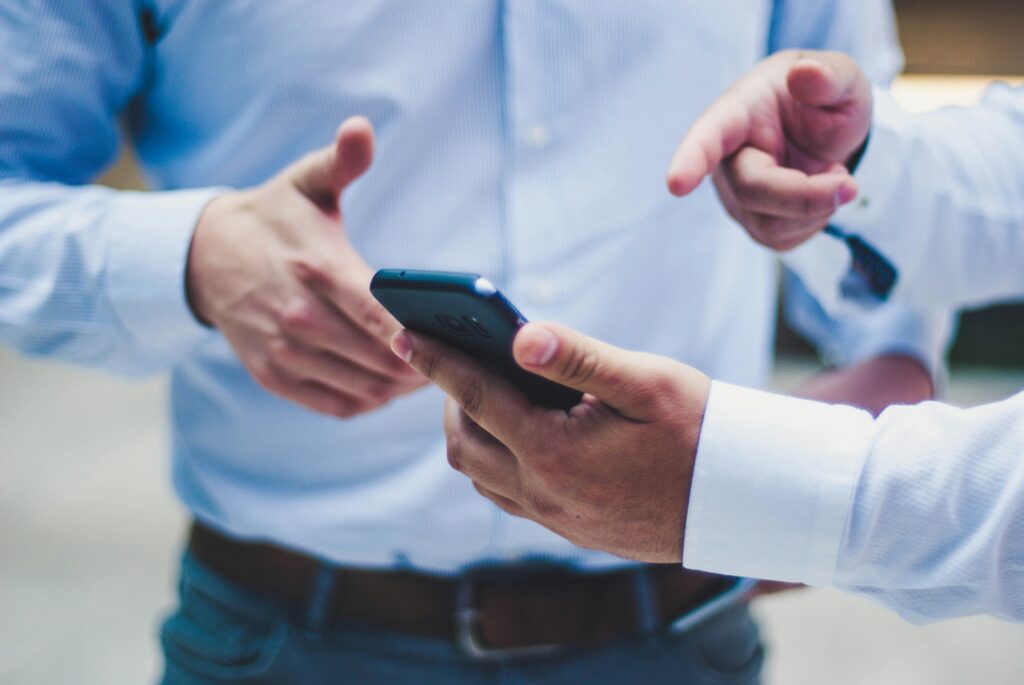 Dois homens conversam enquanto um deles segura um smartphone, apontando com a outra mão para algo que aparece na tela.