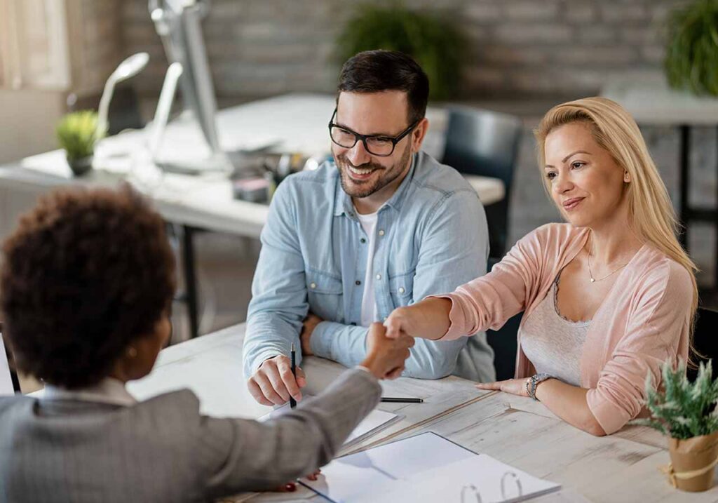 Casal apertando mãos e selando um negócio com uma corretora de imóveis, todos em uma mesa com contratos assinados.